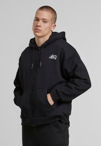 Sweat à capuche noir zippé en coton épais avec une poche frontale, des cordons réglables et un petit logo graphique blanc sur la poitrine gauche.