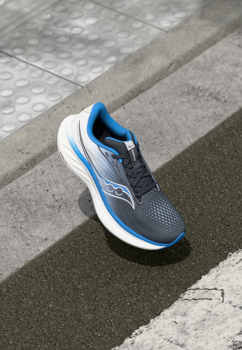 Chaussure de sport grise avec des accents bleus, tige en maille et semelle texturée, positionnée sur un trottoir à côté d'un passage piéton.