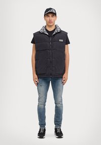 Gilet noir rembourré avec fermeture éclair à l'avant, présentant un col à motifs blanc et noir, associé à un jean bleu déchiré et des baskets noires.
