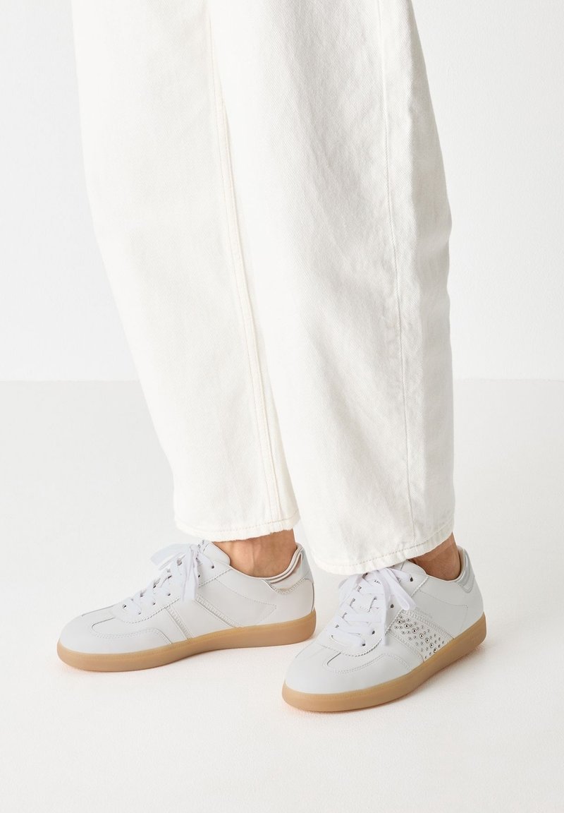 Chaussures de sport gris clair avec des détails perforés sur le côté, des lacets blancs et une semelle en caoutchouc beige, associées à un pantalon blanc ample.