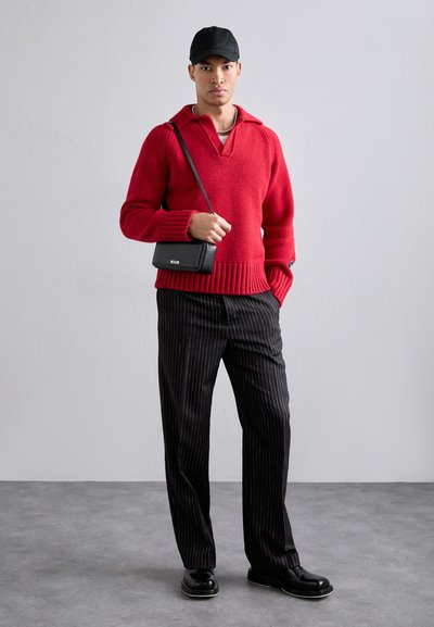 Red knitted pullover with a V-neck and ribbed hem, paired with black pinstriped trousers and black shoes. Accessories include a small black bag.