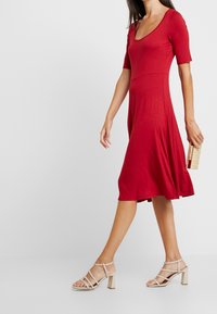 Punainen lyhythihainen mekko, jossa on pyöreä pääntie, väljä A-linjainen leikkaus ja sileä materiaali. Yhdistetty vaaleanbeigeihin hihnakenkiin ja pieneen laukkuun.