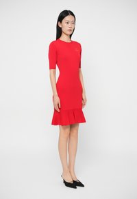 Γυναίκα με μακριά μαύρα μαλλιά, φορώντας ένα εφαρμοστό κόκκινο φόρεμα με μανίκια έως τον αγκώνα και φαρδιά ασύμμετρη φούστα, στέκεται μπροστά σε απλή ανοιχτού χρώματος φόντο.