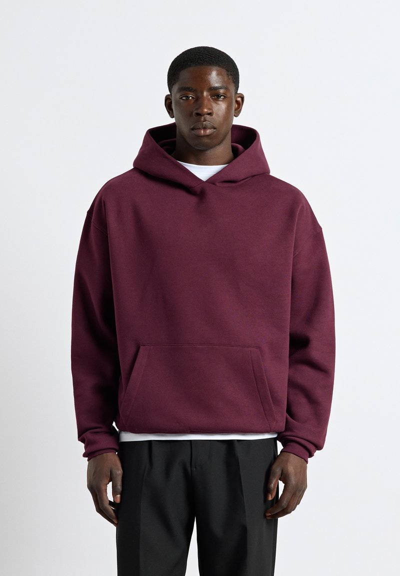 Jeune homme portant un sweat à capuche bordeaux avec poche avant et pantalon noir, debout devant un fond uni clair.