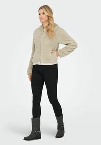 Woman wearing a beige fuzzy zip-up jacket, black slim pants, and black mid-calf boots standing with one hand in pocket.