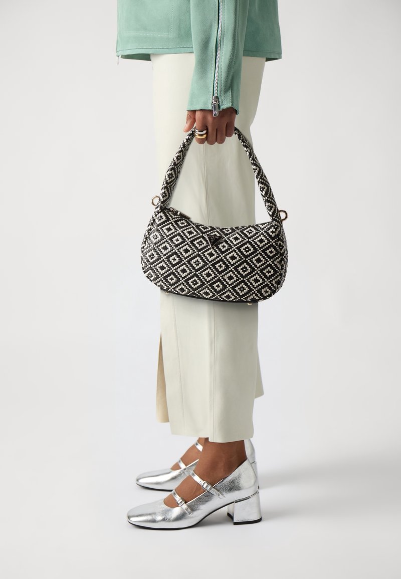Personne portant des talons blocs bas argentés et un pantalon crème tient un sac à main à motifs noir et blanc avec une poignée courte.