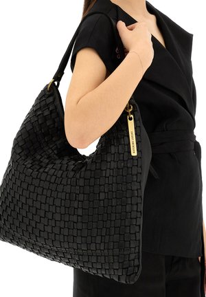 Donna che porta una grande borsa a tracolla in pelle intrecciata nera con targhetta dorata, indossando un vestito nero avvolgente a maniche corte con cintura.
