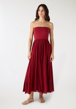 Donna con capelli lunghi e scuri che indossa un abito rosso lungo fino a terra senza spalline e sandali beige, in piedi contro uno sfondo bianco semplice.