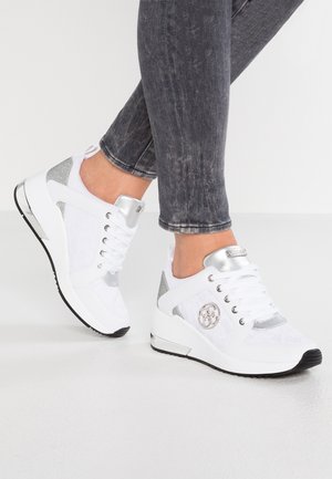 Hvide og sølvfarvede kilehælssneakers båret med mørkegråvaskede skinny jeans, der viser detaljer af skoene fra siden og foran på hvidt gulv.