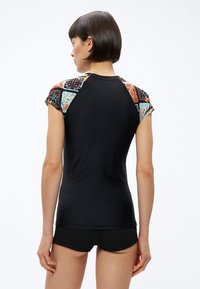 Γυναίκα που φαίνεται από πίσω, φοράει εφαρμοστή μαύρη μπλούζα με πολύχρωμα μανίκια με σχέδια και μαύρα σορτς, μπροστά σε απλό λευκό φόντο.