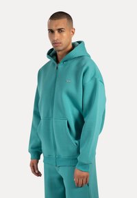Felpa con zip color turchese, dotata di una grande tasca anteriore, polsini a costine e cappuccio regolabile. Realizzata in morbido tessuto testurizzato.