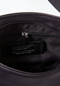 Interiér černé látkové tašky s kapsou na zip; obsahuje plochou štítku s nápisem "United Colors of Benetton." Hladký povrch, žádný viditelný vzor.