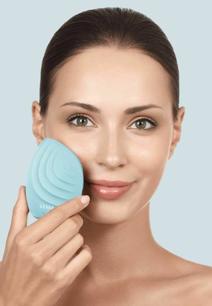 Femme à la peau claire tenant une brosse de nettoyage facial en silicone bleue contre sa joue, souriante sur un fond bleu clair.