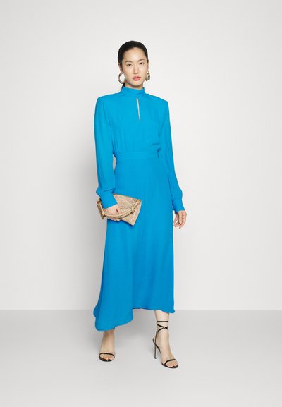 Niebieska sukienka z długim rękawem, z wysoką szyją, detalem w kształcie klucza, wcięciem w talii i luźną spódnicą. Uzupełniona złotą kopertówką i sandałami na obcasie.