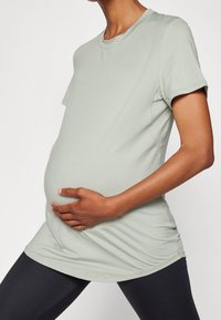 Camiseta de maternidad verde claro con mangas cortas, corte relajado, dobladillo curvado, combinada con leggings negros. Textura suave, diseño sencillo.