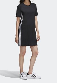 Kobieta ubrana w czarną sukienkę Adidas z białymi paskami po bokach oraz białe trampki Adidas z czarnymi paskami, stojąca na jednolitym tle.