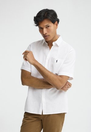 Mand med krydsede arme iført hvid kortærmet skjorte og brune bukser, foran en ensartet lys baggrund.