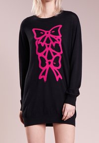 Mustahuta musta mekko, jossa on pitkät hihat ja ribattu pääntie, etupuolella suuri, kirkkaan vaaleanpunainen nauhakuva. Pehmeä tekstuuri.