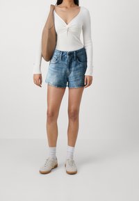 Pantaloni shorts di denim azzurro chiaro con taglio dritto, abbinati a una maglietta bianca a maniche lunghe con dettaglio annodato e una borsa a spalla marrone.