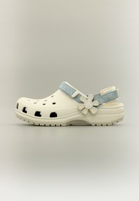 Sabot beige avec des trous d'aération, une bride bleue réglable, un bouton en forme de fleur et une semelle texturée sur un fond neutre.