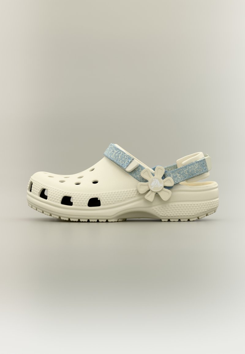 Sabot beige avec des trous d'aération, une bride bleue réglable, un bouton en forme de fleur et une semelle texturée sur un fond neutre.