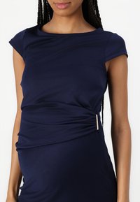 Vestido azul-marinho com mangas curtas, detalhe franzido na lateral e um acento metálico na cintura. Tecido suave com textura lisa.