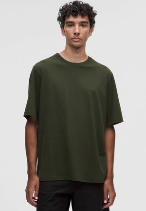 Giovane uomo con capelli ricci e scuri che indossa una t-shirt oversize verde scuro, semplice, a maniche corte su sfondo neutro.