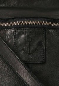 Primo piano di pelle nera con cerniera visibile, logo ancorato in rilievo e dettagli di cucitura. Il testo recita “HARBOUR 2nd.”