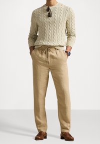Pull en maille beige clair, pantalons en lin beige décontractés avec cordon de serrage et chaussures en cuir marron. Lunettes de soleil reposant sur le pull.