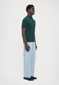 Polo shirt verde închis, cu mâneci scurte, design simplu. Blugi largi, decolorati. Pantofi negri. Model încrucișat spre stânga, pe fundal alb.