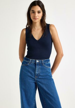 Ung kvinde med langt mørkt hår iført en navyblå ærmeløs V-hals top og højtaljede blå jeans, stående med hænderne bag ryggen.