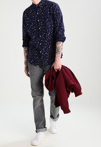 Námořnicky modrá košile na zapínání s bílými vzory, šedé džíny a bílé tenisky; drží vínově červenou bundu; na pažích jsou viditelné tetování.