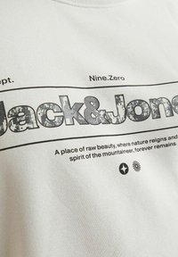Witte sweatshirt met 'Jack & Jones' in textuurrijke, versleten zwarte letters, met aanvullende afgedrukte tekst en grafische accenten eronder.