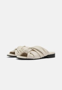 Sandali slip-on in pelle color crema con design a strisce attorcigliate, suola piatta nera e texture liscia.