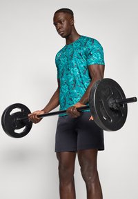 Homme soulevant une barre avec des disques de poids noirs, portant une chemise turquoise à motifs et un short noir, se tenant devant un fond neutre.