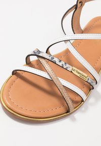 Sandalia con tiras en blanco y dorado, con una suela marrón. Presenta tiras texturizadas en acabados metálicos, de estampado animal y lisos. Accesorios en dorado.