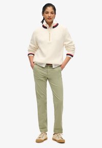 Pull en maille blanche avec une fermeture éclair, col rayé rouge et bleu. Pantalons vert clair et baskets beiges avec une semelle texturée.