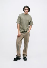 Olivově zelené oversized tričko, volný střih, spárováno se světle béžovými kalhotami s mírným ošetřením, doplněné černými robustními botami.