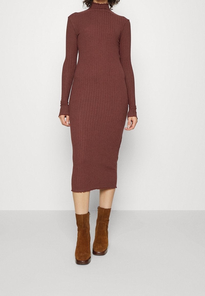 Femme portant une robe midi marron côtelée ajustée à manches longues et des bottines en daim marron, debout devant un fond blanc uni.