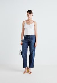 White lace-trimmed camisole with button detail, paired with high-waisted, straight-leg blue denim jeans. Light yellow heeled sandals and small silver handbag.