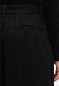 Μαύρο παντελόνι με ρίγες, με πίσω τσέπη welt, φορεμένο με μαύρο ριμπ πλεκτό πουλόβερ, με μέρος ενός χεριού να φαίνεται στη δεξιά πλευρά.