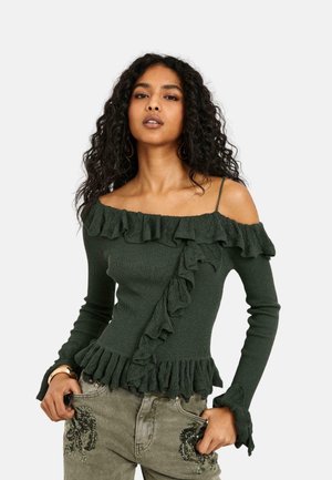 Mujer con cabello largo y rizado que lleva un suéter verde oscuro con volantes y hombros descubiertos y pantalones verde oliva con bordado floral con cuentas.