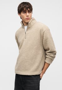 Μπεζ φλις μπλούζα με ψηλό γιακά και ημιδιακοπή με φερμουάρ. Μαλακή υφή, άνετη εφαρμογή και γυρισμένα μανίκια πάνω από σκούρο τζιν.