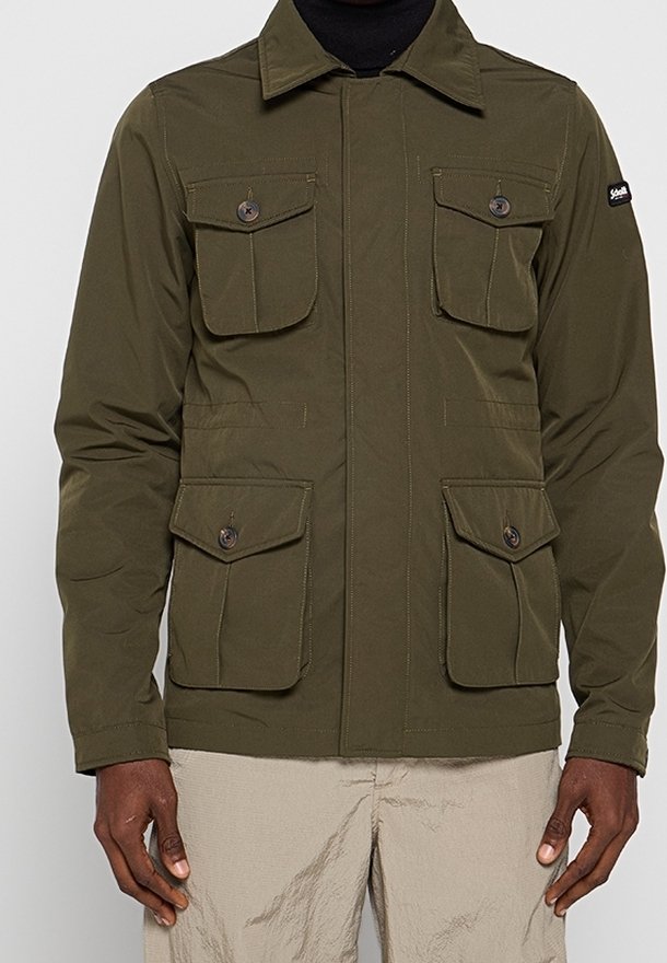 Olivengrøn jakke med fire knappebeslåede lommer foran, båret over en sort top, kombineret med lyse beige bukser.