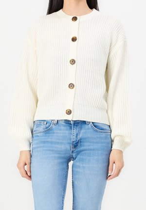 Kvinde iført en hvid strikket cardigan med store brune knapper sammen med lyseblå jeans, stående mod en ensfarvet baggrund.