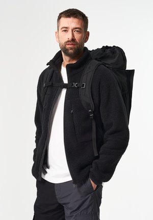 Bearded man wearing black fleece jacket, white T-shirt, and dark cargo pants with black backpack straps and hands in pockets, standing on white background.