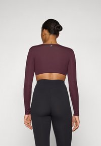 Long-sleeve, cropped top in burgundy stretchy material. Fitted design with a round neckline. Worn with high-waisted black leggings.