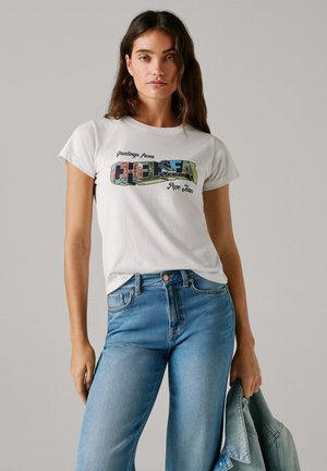 Biele bavlnené tričko s krátkym rukávom, na ktorom je farebná grafika s nápisom "Pozdravy z Chelsea". Nosené s modrými džínsami a v ruke má rifľovú bundu.