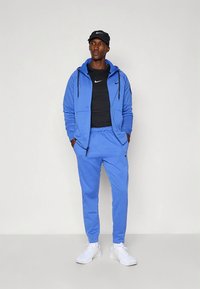 Fato de treino atlético azul com capuz com fecho éclair e calças com cordão, feito de material liso; combinado com uma t-shirt preta e ténis brancos.