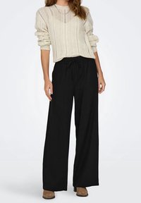 Jasnobeżowy sweter z dzianiny o prążkowanej fakturze, noszony z czarnymi spodniami z szerokimi nogawkami i brązowymi butami, przedstawiony na neutralnym tle.